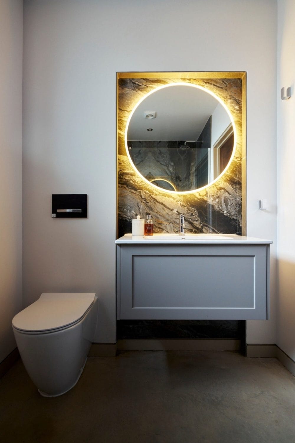 There's a Creative reflection around the LED mirror lighting up the natural stone-effect tiles, with grey vanity and basin along with grey floor tiles and white walls