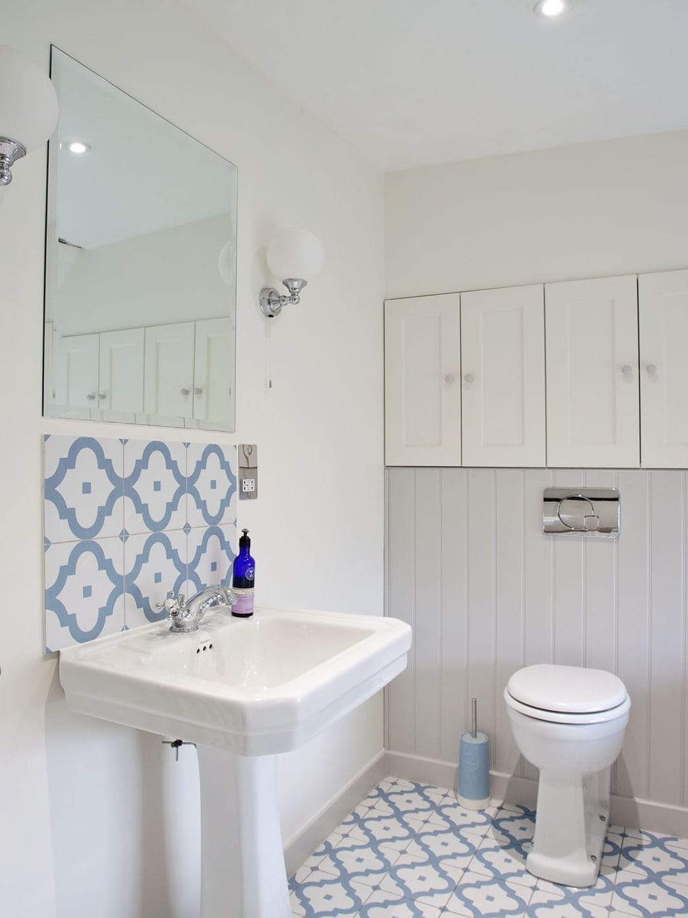 A Bright white bathroom with blue patterned floor tiles, pedestal sink, and shaker cabinets. Showing how light can bounce across reflective surfaces