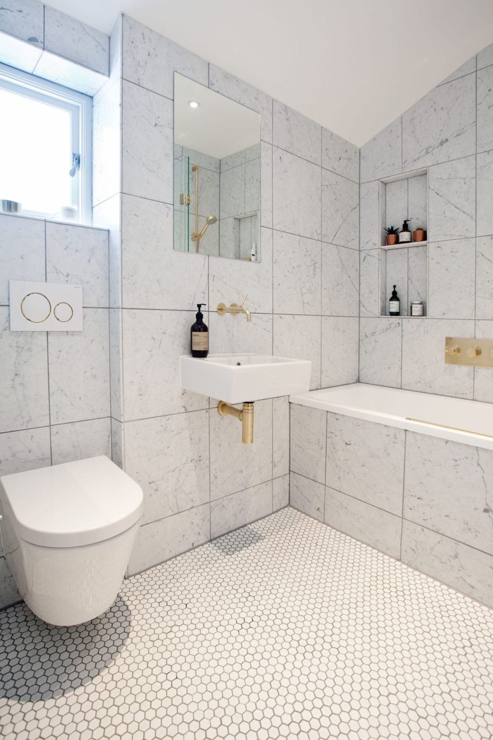 A Modern family bathroom with marble tiles, gold fixtures for a luxury feel, and hexagon floor tiles. Marble sled niches provides minimalistic storage