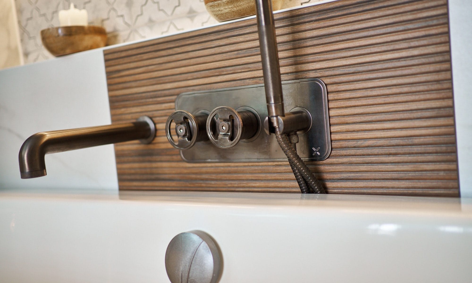 Close‑up of brushed bronze wall‑mounted bath spout and controls set into ribbed wood panel with tiled niche above.