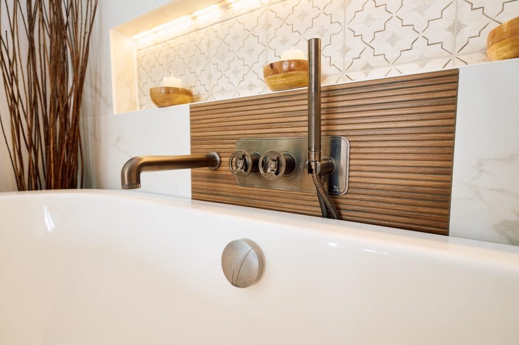 Bronze bath controls and handset mounted on ribbed wood panel below a backlit patterned niche.
