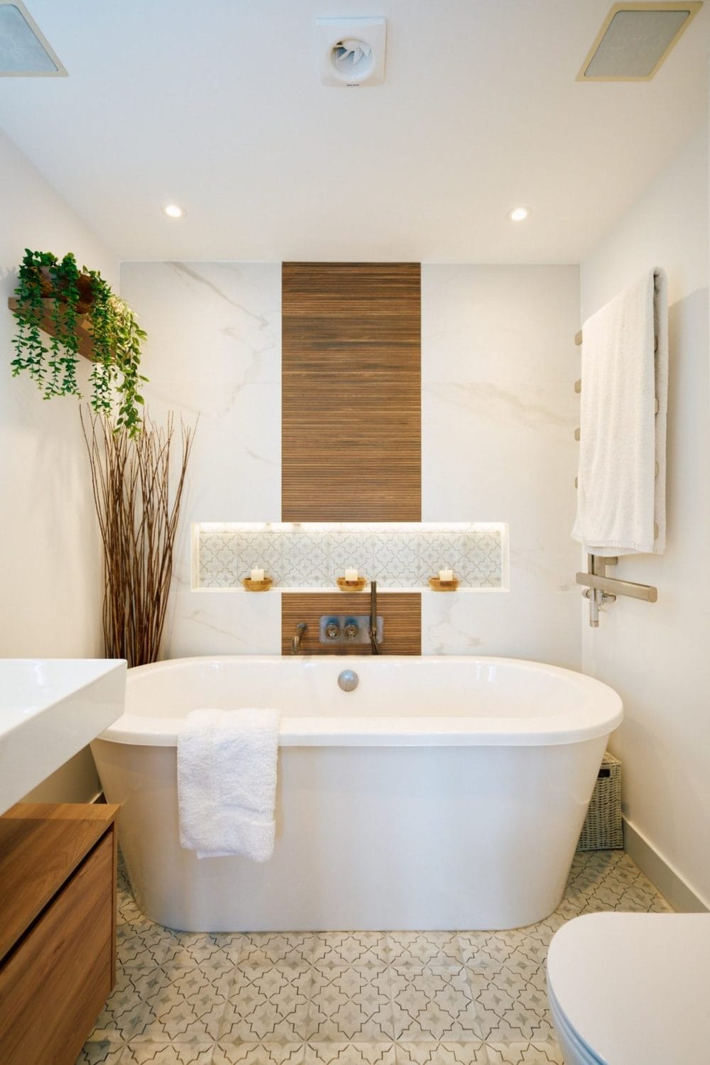 Freestanding white bath with vertical wood‑slat feature panel, backlit niche and marble‑effect wall tiles in a modern Sussex bathroom.