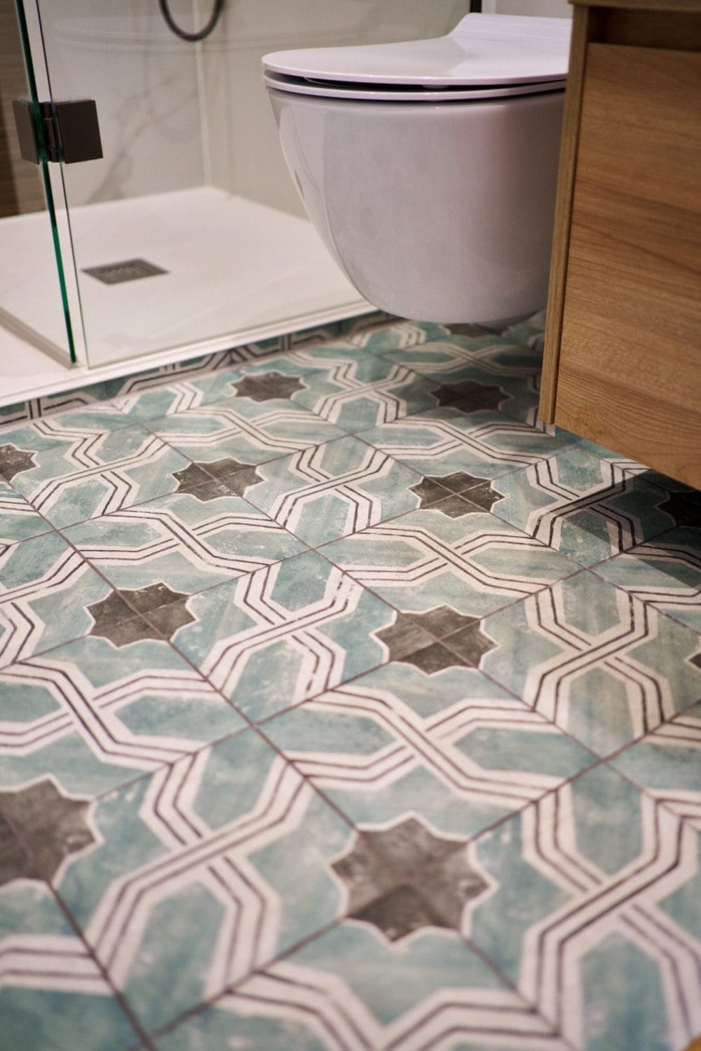 Patterned teal, grey and white floor tiles beneath a wall‑hung WC and frameless shower in a modern bathroom.