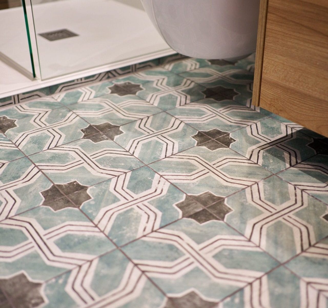 Patterned teal, grey and white floor tiles beneath a wall‑hung WC and frameless shower in a modern bathroom.